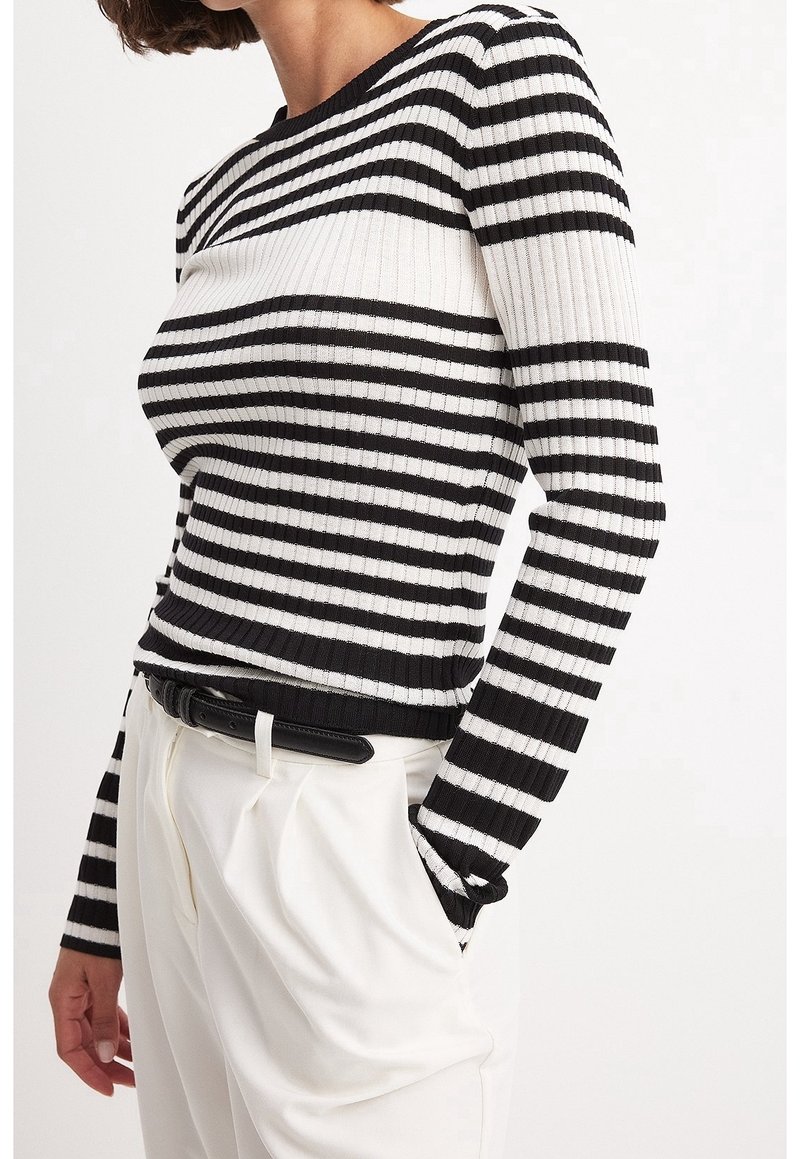 Femme portant un haut à manches longues côtelé à rayures horizontales noires et blanches, rentré dans un pantalon blanc taille haute avec une ceinture noire.