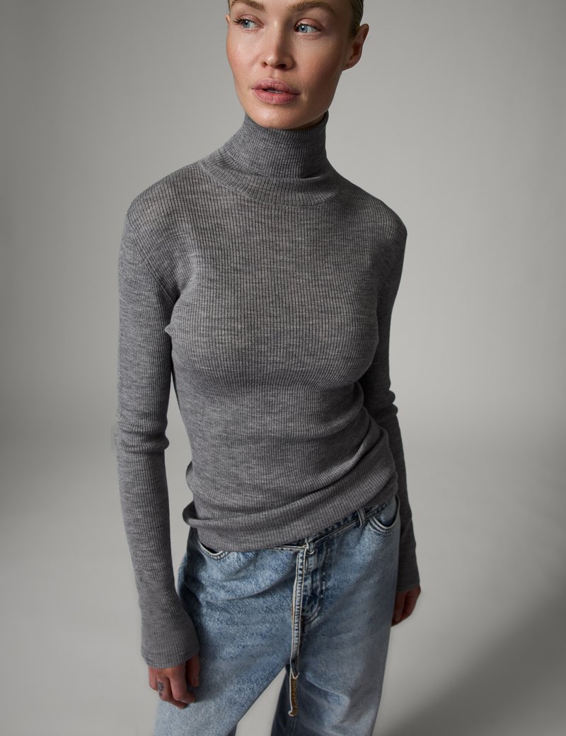 Vrouw draagt een grijze geribde coltrui en lichtblauwe spijkerbroek, kijkt naar links tegen een effen grijze achtergrond.