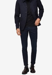 Chaqueta navy a rayas con dos botones, llevada sobre una camiseta negra. Unos vaqueros oscuros de corte ajustado y unos zapatos de cuero negro brillantes completan el look.