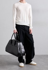 Weißes Langarmshirt und schwarze Cargo-Hosen, getragen von einer Person, die eine schwarze Leder-Reisetasche hält. Die Turnschuhe sind weiß mit beigen Sohlen.