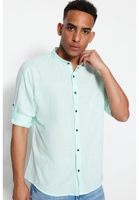 Camicia mint green a maniche corte con colletto alla coreana, caratterizzata da bottoni neri e un tessuto texture, indossata con un gesto casual della mano.