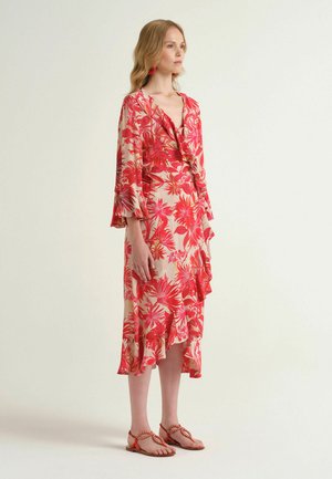 Donna in piedi che indossa un vestito midi color crema con stampa floreale rossa, orlo arricciato, maniche svasate, abbinato a sandali intrecciati corallo e orecchini coordinati.