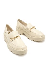 Cream leather loafers featuring a chunky heel, rounded toe, and a decorative gold chain across the front. Textured surface adds detail.