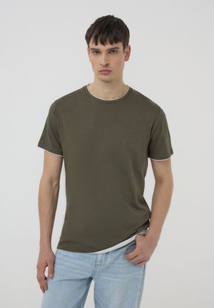 T-shirt verde oliva a maniche corte con rifiniture bianche sul collo e sulle maniche, realizzata in tessuto leggero, indossata con jeans azzurri chiari.
