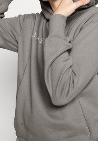 Graue Kapuzenjacke aus weichem Material, mit einer Vordertasche, gerippten Bündchen und einer Stickerei auf der Brust. Sanfte Textur, entspannte Passform.