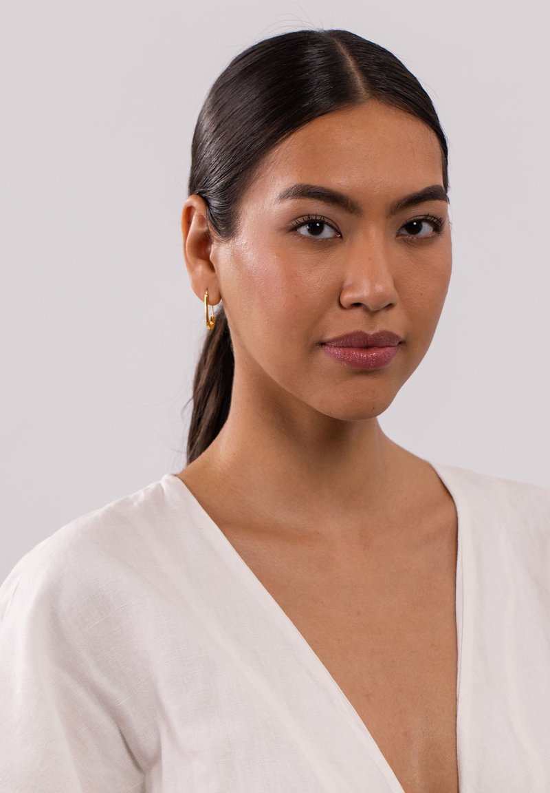Model trägt ein weißes Leinenoberteil mit tiefem V-Ausschnitt und dünnen Trägern, dazu kleine goldene Creolen und glattes schwarzes Haar.