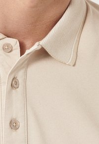 Beige polo skjorta med ett strukturerat tyg, med en standardkrage och tre knappar i matchande färg. Diskret accentstickning.