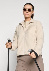 Chaqueta de forro polar color beige claro con cierre frontal y superficie texturizada. Llevada sobre una camisa color crema, el modelo sostiene bastones de senderismo negros.