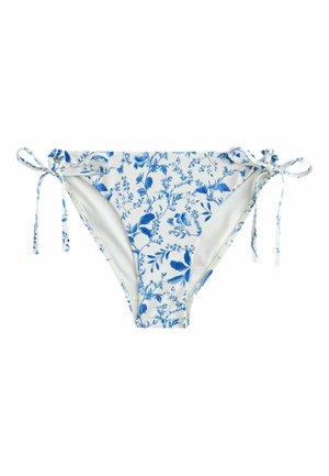 Lächelnde Frau mit welligem blondem Haar, die einen weißen Bikini mit Blumenmuster trägt und sich an eine weiße Wand am Meer bei klarem Himmel lehnt.