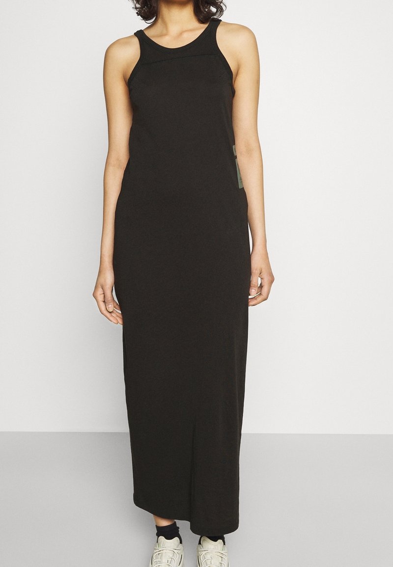 Femme portant une robe maxi noire sans manches associée à des baskets blanches, debout contre un mur et un sol gris clair unis.