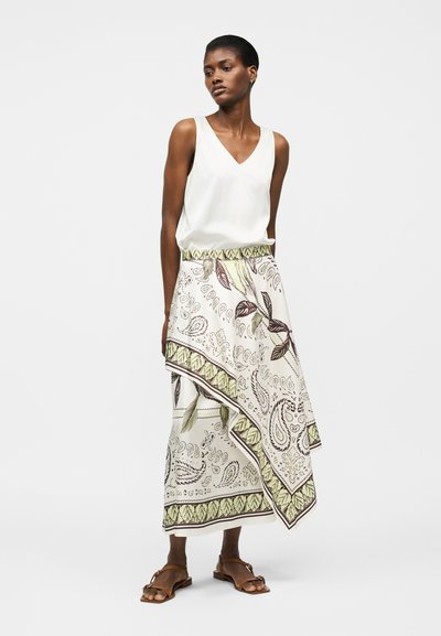 Γυναίκα που στέκεται φορώντας λευκή αμάνικη μπλούζα με V-λαιμόκοψη, ασύμμετρη φούστα με σχέδια φύλλων και καφέ φλατ σανδάλια σε λευκό φόντο.