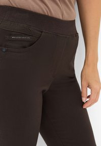 Brune strækbukser har en bred talje, sidelomme med metalaccent og glat tekstur. Tæt på visning af talje og hånd.