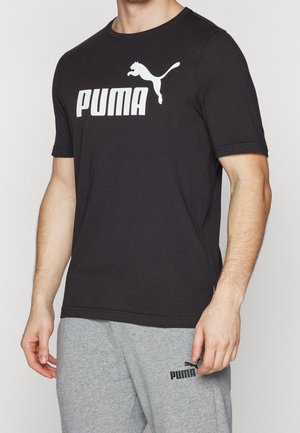 Mężczyzna w czarnej koszulce Puma i jasnoszarych spodniach dresowych Puma stoi na jednolitym tle.