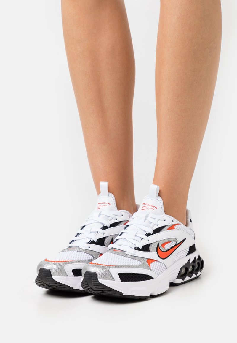 Baskets de sport blanches avec des accents noirs et orange, dessus en maille, semelle épaisse et design de talon distinct. Les lacets sont soigneusement noués.