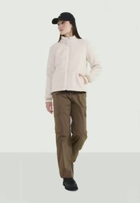 Giacca in pile rosa chiaro con zip frontale, abbinata a pantaloni cargo marroni larghi e calzature nere. Accessoriata con un cappellino nero.
