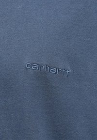 Modrý textil s vyšívaným logem Carhartt ve shodné modré nitě na hladkém textilním povrchu.