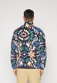 Chaqueta de forro polar con cuello alto, con un patrón floral colorido en azul, verde, durazno y blanco roto sobre un fondo oscuro.