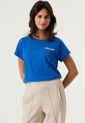 Blauw katoenen T-shirt met opgerolde mouwen en een geborduurd "Amore" in goud. Gecombineerd met licht beige broek, getextureerd met subtiele details.