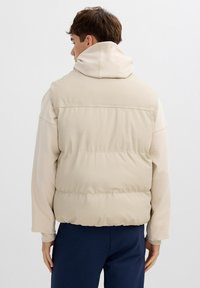 Chaleco acolchado beige con cuello alto, acabado liso y costuras laterales. Lleva encima una camiseta de manga larga beige claro, combinando con pantalones azul marino.