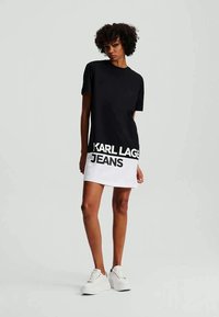 Vestido t-shirt preto com bainha branca e texto "KARL LAGER JEANS". Mangas curtas, design linear, tecido suave, combinado com sapatilhas brancas.