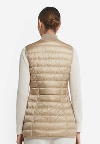Beige vadderad väst med hög krage, vertikal quiltad design och ribbade ärmhål, bärs över en ljusfärgad tröja.