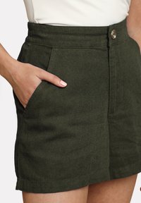 Olivengrøn kort nederdel med en glat tekstur, høj talje, frontknaplukning og sidelommer. Enkel, minimalistisk design.