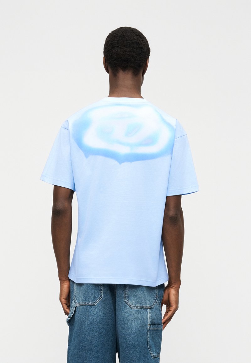 Άνδρας στέκεται με την πλάτη, φοράει ανοιχτομπλε μπλουζάκι με λευκό και μπλε κυκλικό σύννεφο στο πάνω μέρος της πλάτης και τζιν σορτς.