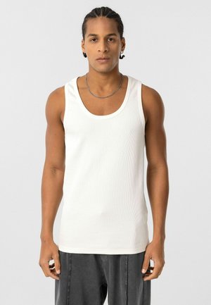 Witte geribbelde tanktop met een ronde hals. Soepele textuur, aansluitende pasvorm en brede schouderbanden. Draagt met donkere broeken.