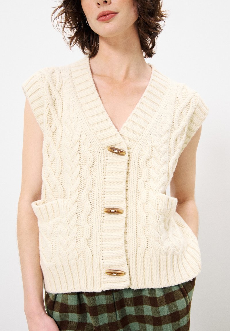 Cremfarbener Strickweste mit V-Ausschnitt, dekorativen Holzknöpfen und Seitentaschen. Mit gerippten Kanten und kurzen Ärmeln.