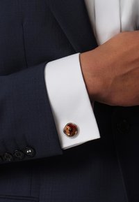 Cufflink featuring a round, amber-colored stone set in silver, attached to a white dress shirt with a navy blazer and black buttons.
