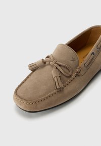 Beige mockasin-sko i mocka med rund tå, dekorativa fransar och kontrasterande sömmar. Har snörhål och texturerad sula.