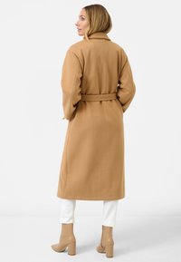 Cappotto lungo in cammello con un finish testurizzato, vita cintata, collo a rever e maniche ampie, abbinato a pantaloni bianchi e stivaletti beige alla caviglia.