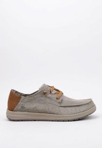 Mocasin beige de hombre casual con parche de talón marrón claro, cordones color beige, parte superior de tela y suela de goma, mostrado en perfil lateral sobre fondo blanco.