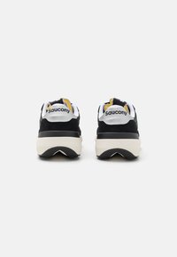 Zapatillas deportivas en blanco y negro con una parte superior texturizada, que cuentan con acentos amarillos y una suela de goma resistente, mostrando la marca "Saucony."