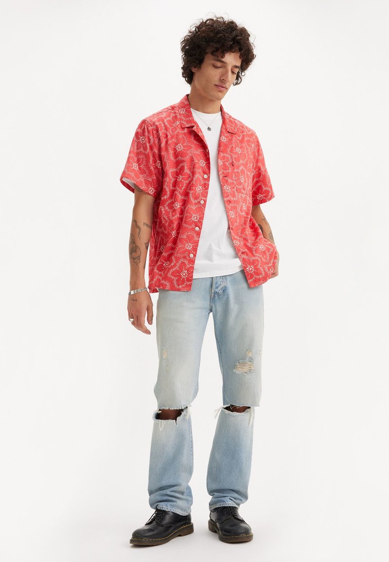 Camicia rossa a maniche corte con motivo floreale, indossata sopra una maglietta bianca, abbinata a jeans strappati blu chiaro e scarpe nere con lacci.
