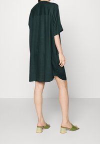Tunika dress verde scuro, morbida e a maniche corte, con scollatura rotonda e orlo curvo. Abbinata a mules verdi con punta aperta e tacco a blocco.