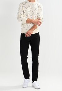 Maglione lavorato a maglia di colore crema con un motivo a diamante, abbinato a jeans neri slim-fit e sneakers bianche. Il tessuto appare texturizzato.