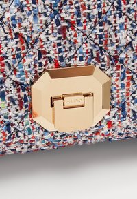 Fermoir géométrique ton doré sur tissu tissé multicolore avec des fils bleus, rouges, blancs et noirs, comprenant un motif matelassé cousu.