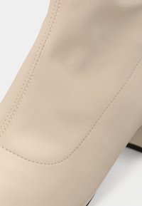 Prim-plan al unei cizme din piele bej, cu cusături evidente și vârf pătrat, evidențiind textura netedă și cutele subtile.