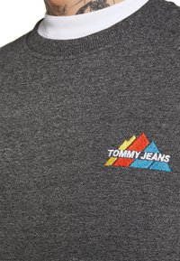 Šedá mikina s kulatým výstřihem, vyrobená z texturované látky a zdobená barevným vyšívaným logem v červené, žluté a modré barvě.