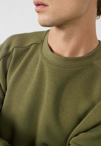Primer plano de una persona que lleva un sudadera de cuello redondo en verde oliva, mostrando el cuello, la línea de la mandíbula y la parte superior del torso.