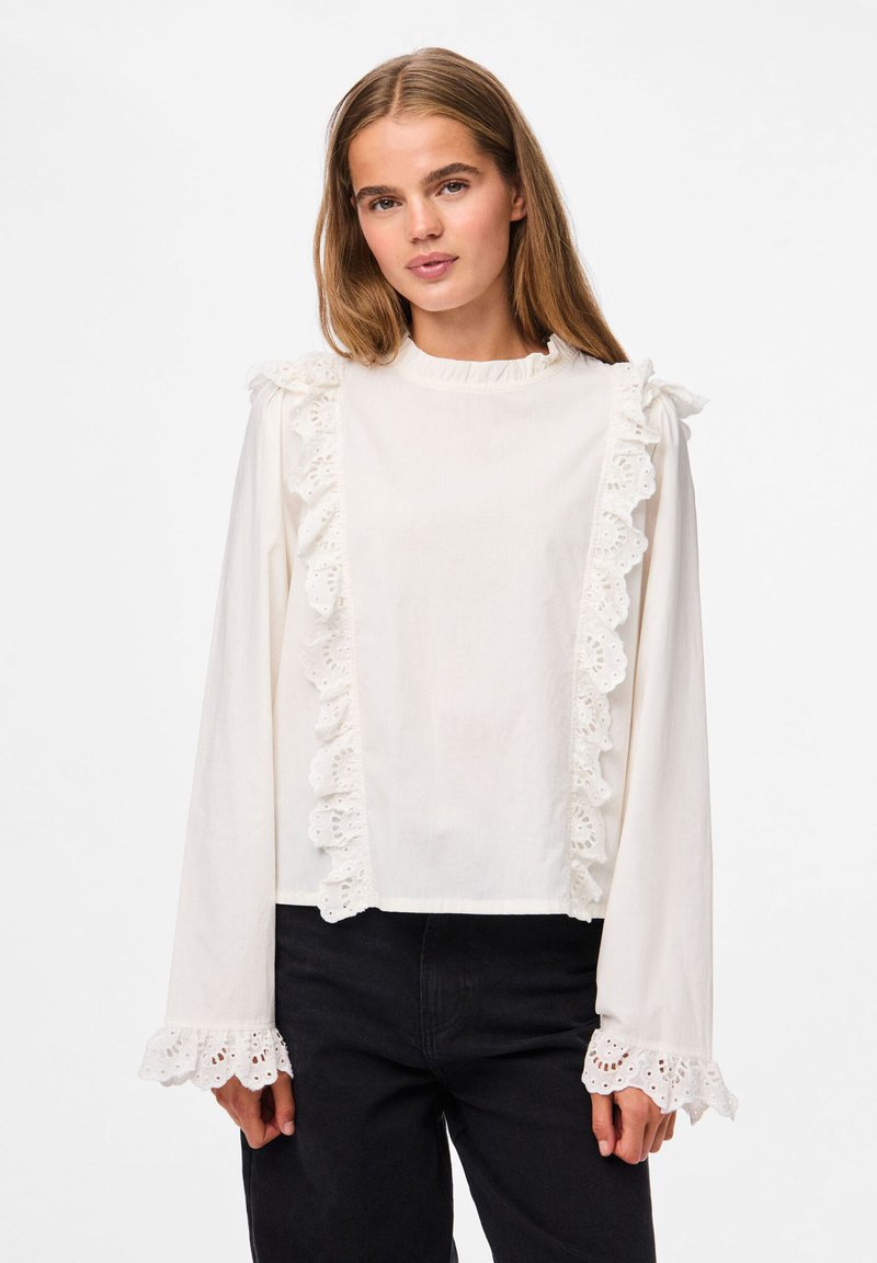 Blusa blanca de manga larga con volante de encaje a lo largo del escote y las mangas, con corte recto y fabricada en un tejido de algodón ligero.