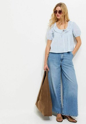 Młoda kobieta w okularach przeciwsłonecznych, jasnoniebieskiej bluzce z bufkami, szerokich dżinsach, brązowych sandałach, trzymająca dużą brązową torbę na ramię, stoi na tle białej ściany.