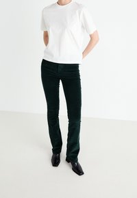 T-shirt en coton blanc associé à un pantalon en velours côtelé vert, avec une coupe ajustée. Des bottines à talons noires à bout carré complètent la tenue.