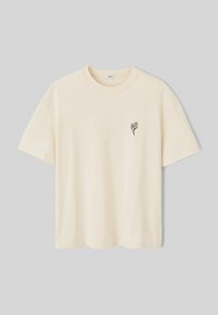 T-shirt en coton crème avec une coupe décontractée, des manches courtes et une broderie florale noire sur la partie gauche de la poitrine. Texture lisse.