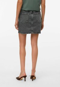 Mini-jupe en denim noir au design ajusté, avec cinq poches et un ourlet effilé. Portée avec des mules à talons bruns et un haut vert ajusté.