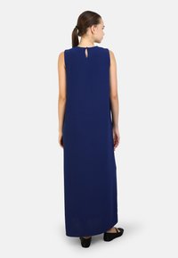 Abito maxi blu navy con design senza maniche e dettaglio chiave sulla schiena, realizzato in tessuto morbido. Indossato con scarpe basse nere.