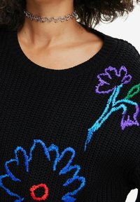 Schwarzer Strickpullover mit buntem Blumenstickerei in Blau, Rot und Grün. Choker-Halskette mit silbernem, verflochtenem Design.