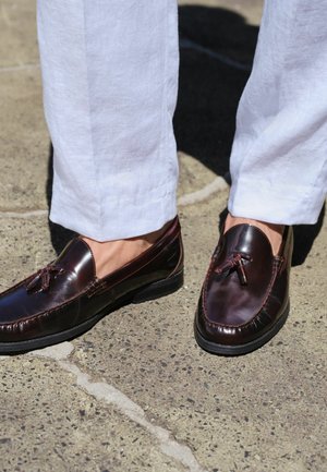 Dark brown leather tassel loafers worn with white linen trousers, standing on a textured concrete surface.