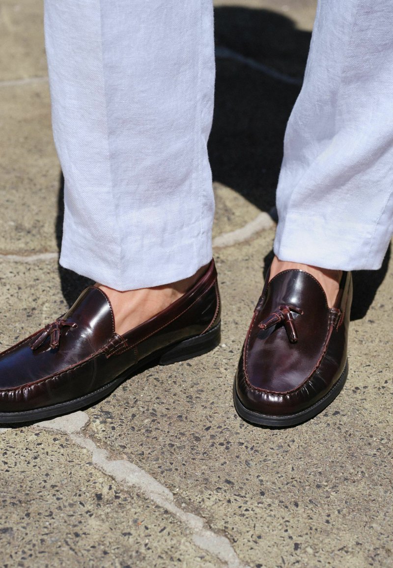 Mocassins à glands en cuir marron foncé portés avec un pantalon en lin blanc, debout sur une surface en béton texturée.